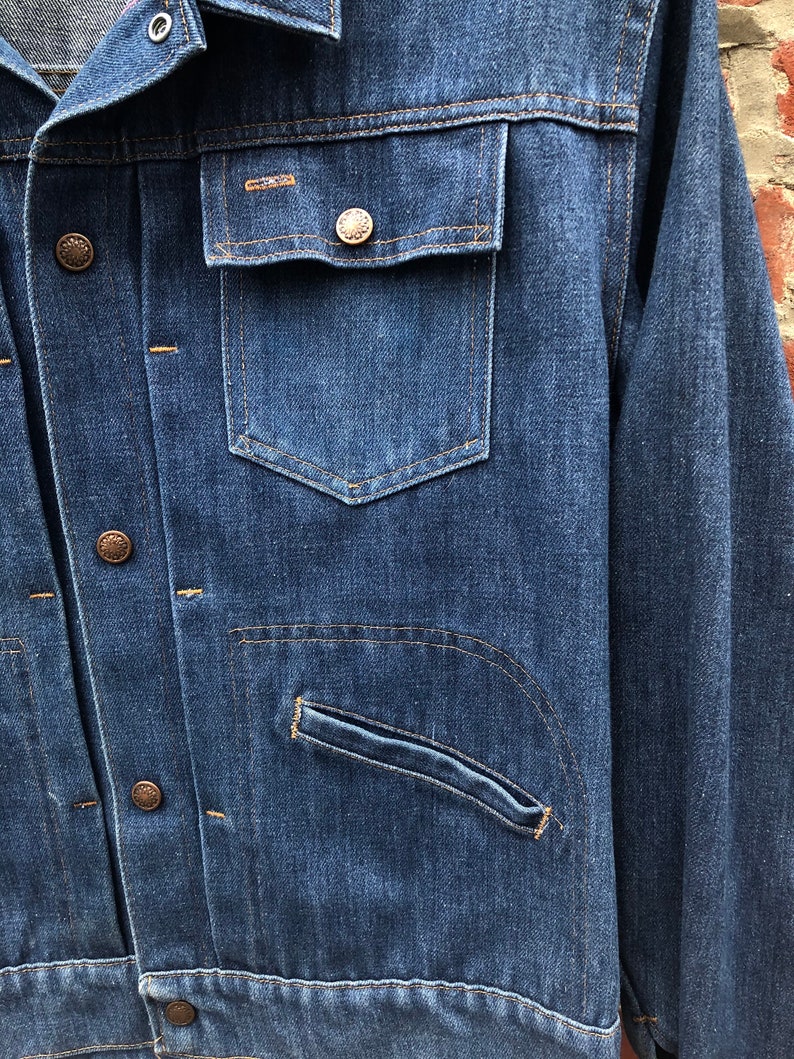 May include: A blue denim jacket with a button closure, two chest pockets, and a single pocket on the left sleeve. The jacket has a worn look and is made of a thick, durable fabric.