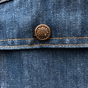 May include: Close-up of a blue denim jacket with a copper-colored button. The button has a circular design with a starburst pattern.