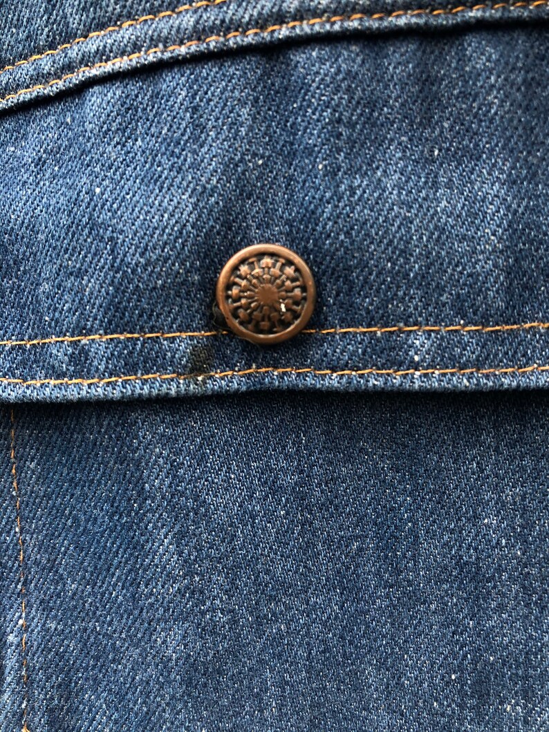 May include: Close-up of a blue denim jacket with a copper-colored button. The button has a circular design with a starburst pattern.