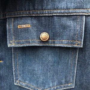 May include: A close-up of a blue denim jacket with a single pocket and a gold button. The pocket has a contrasting orange stitch.