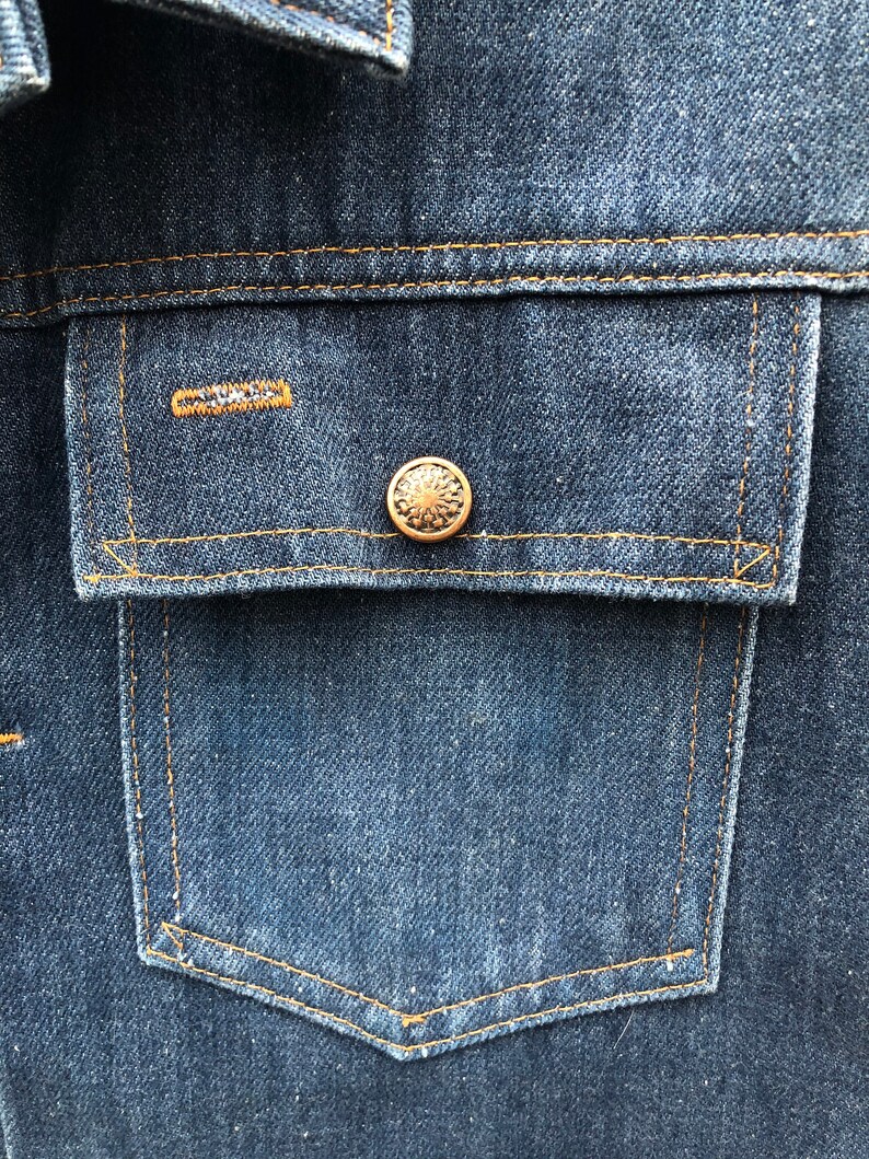 May include: A close-up of a blue denim jacket with a single pocket and a gold button. The pocket has a contrasting orange stitch.