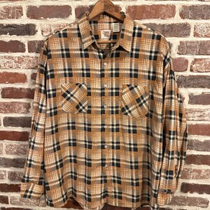 May include: A long-sleeved plaid shirt with a brown, black, and white plaid pattern. The shirt has a button-down collar and two chest pockets. The shirt is hanging on a wooden hanger.