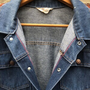 May include: A blue denim jacket with a red stitching detail on the inside of the collar. The jacket has a button closure and two flap pockets on the chest. The label on the inside of the jacket reads "Indigo Super Denim".
