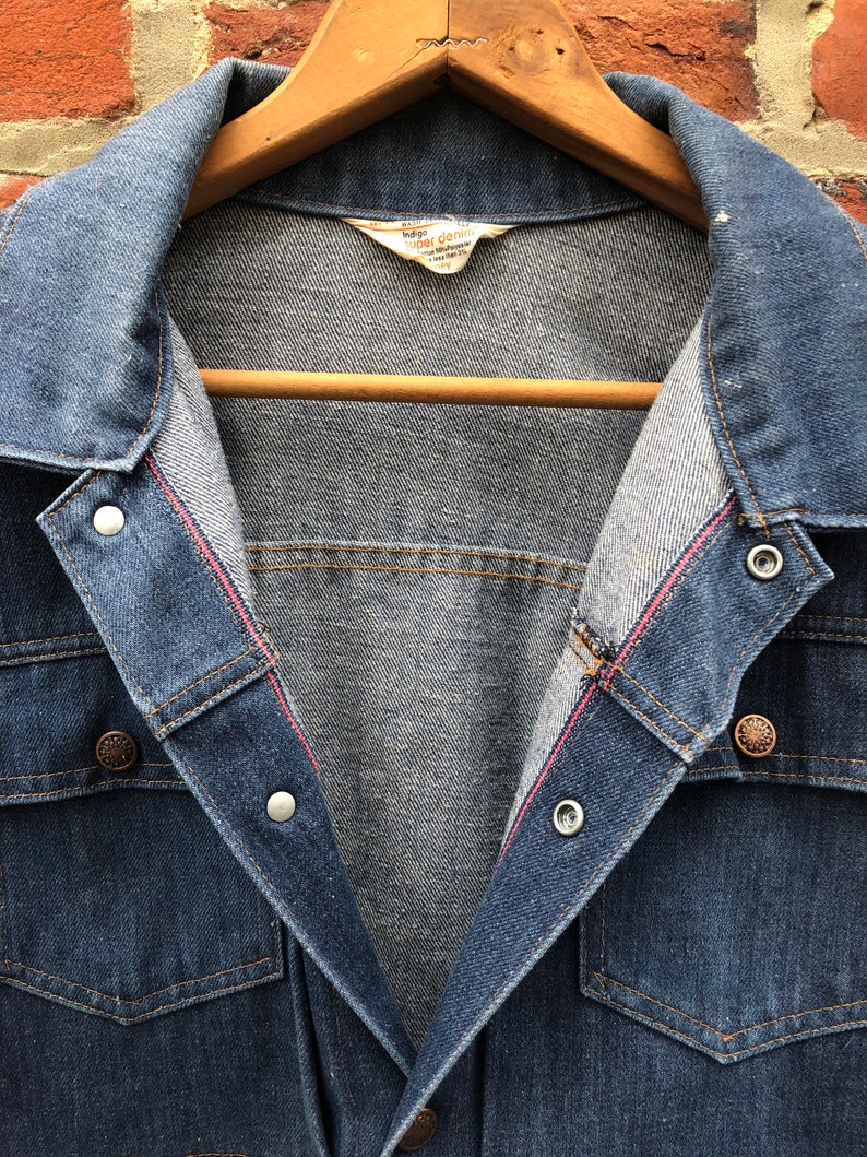 May include: A blue denim jacket with a red stitching detail on the inside of the collar. The jacket has a button closure and two flap pockets on the chest. The label on the inside of the jacket reads "Indigo Super Denim".