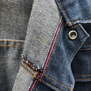 May include: Close-up of a denim jacket with a red stitching detail. The inside of the jacket is a lighter shade of denim with a white stripe pattern.