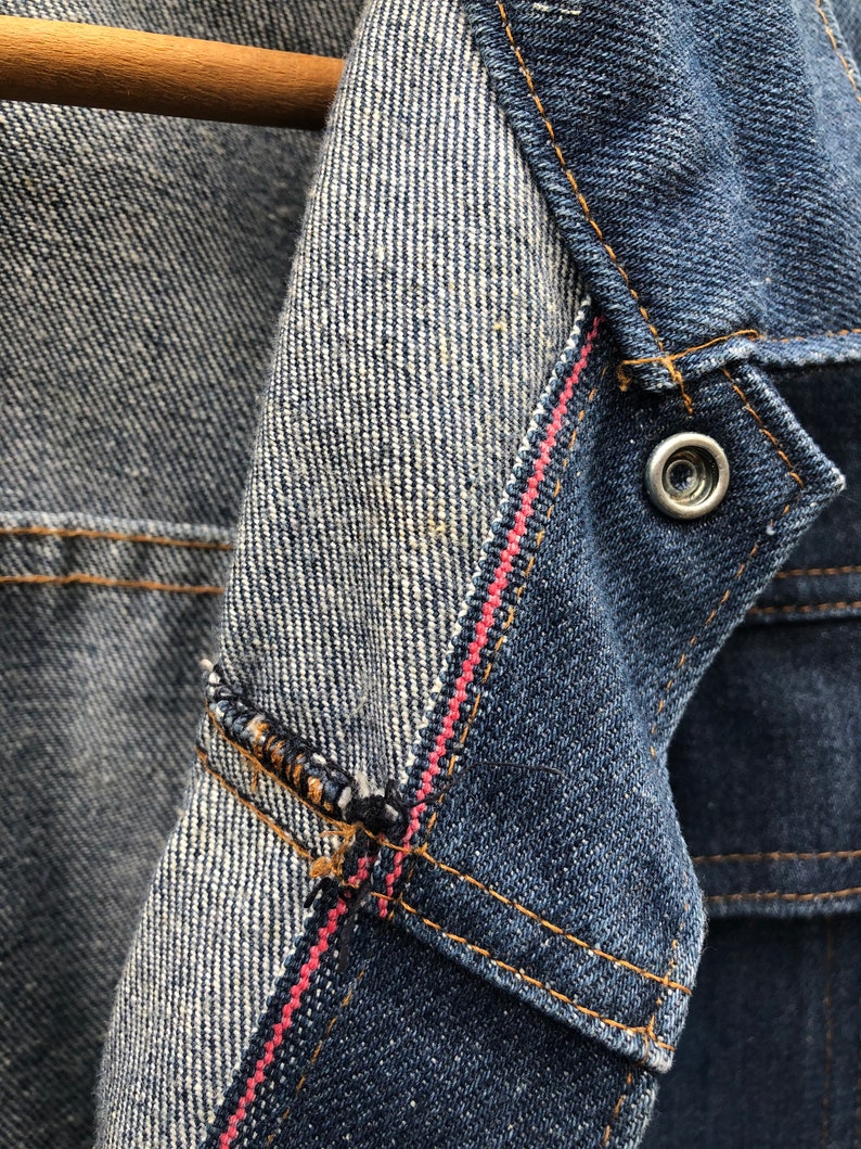May include: Close-up of a denim jacket with a red stitching detail. The inside of the jacket is a lighter shade of denim with a white stripe pattern.