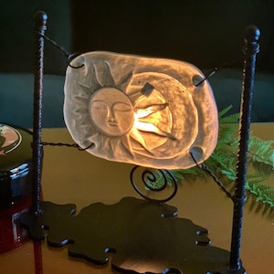 May include: A black metal stand with a sun and moon design. The sun is made of glass and is lit from behind. The stand is on a dark red surface.