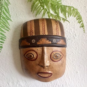 May include: A tan-colored, carved mask with a stylized face. The mask features a brown hat with vertical stripes and a dark brown band. The face has almond-shaped eyes and a small, open mouth. Decorative elements include circular and geometric patterns.