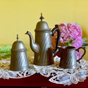 May include: A silver tea set with a teapot, creamer, and sugar bowl. The set is decorated with a swirl pattern and sits on a white lace doily.