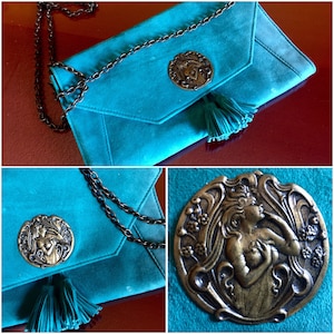 May include: A teal blue suede clutch bag with a metal chain strap and a decorative metal clasp featuring a woman's face. The bag has a tassel hanging from the clasp.