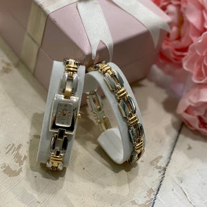 May include: Two gold and silver tone bracelets with a watch on one of the bracelets. The bracelets are on white display stands. The bracelets are next to a pink gift box with a white bow.