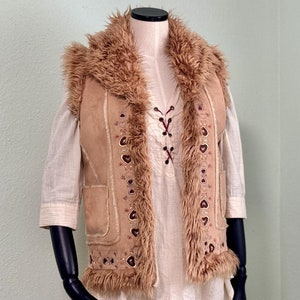 Op de afbeelding: Een bruin nep bontvest met suède accenten en geborduurde bloemdetails. Het vest heeft een nep bontkraag en een vetersluiting aan de voorkant.