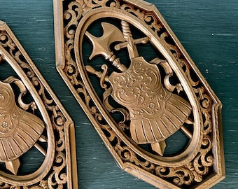 May include: Two ornate gold-coloured wall plaques with a decorative design featuring a shield, sword, and axe. The plaques are shaped like a shield and have a raised, detailed design.