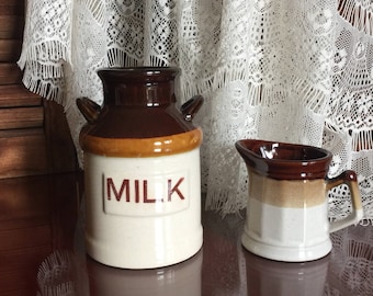 Könnte beinhalten: Eine braune und weiße Keramik-Milchkanne mit dem Wort "MILK" auf der Vorderseite und eine kleinere braune und weiße Keramik-Sahnegießkanne daneben.