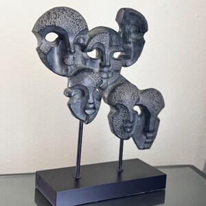 May include: A black abstract sculpture of multiple faces on a black base. The sculpture is made of a textured material and has a modern design.