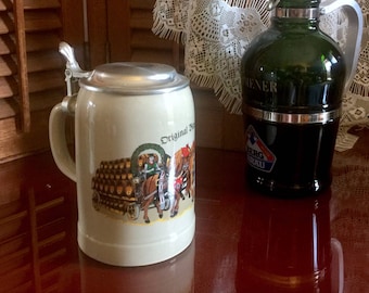 May include: A white ceramic beer stein with a pewter lid and a scene of a horse-drawn wagon carrying barrels of beer. The stein has the text "Original Wiener" printed on it. A green glass bottle with a silver lid and the text "Original Wiener" is also in the image.