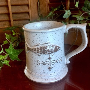 May include: A white ceramic mug with a brown fish design and a compass rose. The mug has a speckled glaze and a brown handle.