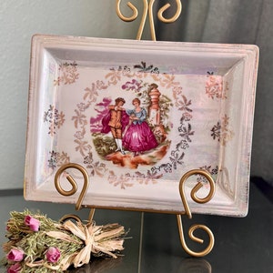 May include: A white porcelain tray with a gold trim and a romantic scene of a couple dancing in a garden. The tray is on a gold metal stand.