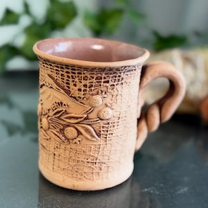 Könnte beinhalten: Eine braune Keramiktasse mit einer strukturierten Oberfläche und einem Reliefmuster aus Blättern und Beeren. Die Tasse hat einen gedrehten Henkel.