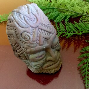 May include: A brown ceramic tiki mask with intricate carvings and a closed-eye expression. The mask is decorated with a geometric pattern and a swirl design.