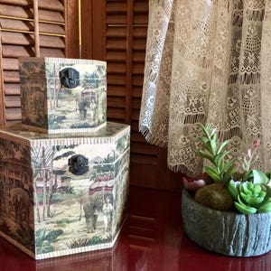 May include: Two hexagonal decorative boxes with a vintage elephant and landscape design. The boxes are stacked, with a smaller one on top of a larger one. Each box has a black metal clasp. A small succulent arrangement sits nearby.