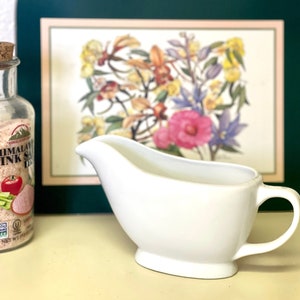 May include: A white ceramic gravy boat with a handle. The boat is sitting on a light brown surface in front of a framed picture of flowers.