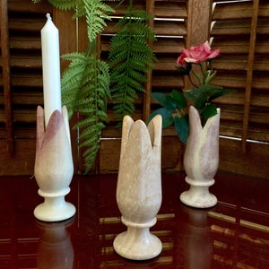 Puede incluir: Tres candelabros decorativos en forma de tulipán hechos de mármol blanco y rosa. Los candelabros están sobre una superficie roja. Un candelabro tiene una vela blanca dentro.