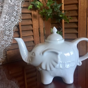 May include: White ceramic teapot shaped like an elephant. The teapot has a lid and a spout. The teapot is sitting on a wooden surface.
