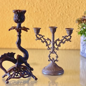 May include: Two ornate metal candle holders. One is a three-armed candelabra with a scrolled design. The other is a single-armed holder shaped like a dragon.