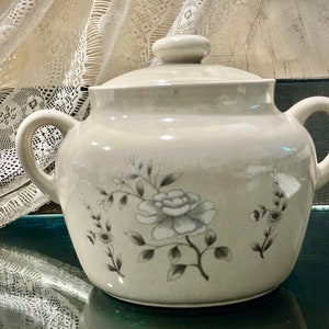 May include: A white ceramic crock with a lid, featuring a floral design in shades of gray. The crock has two handles and a lid with a knob.
