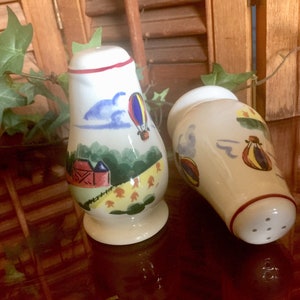 May include: A pair of ceramic salt and pepper shakers with a whimsical hot air balloon theme. The shakers are painted with a white background and feature colorful hot air balloons, clouds, and a red barn.
