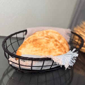 May include: Two round loaves of bread in a black wire basket lined with a light blue and beige fabric. The basket is on a dark surface.