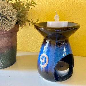 Puede incluir: Un quemador de aceite de cerámica azul y marrón con un diseño de espiral dorado. El quemador tiene una pequeña abertura para una vela de té y una abertura más grande para aceites esenciales.