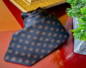 Könnte beinhalten: Eine marineblaue Krawatte mit einem sich wiederholenden Muster aus kleinen, goldfarbenen Kreisen. Die Krawatte ist aus glattem Stoff gefertigt und wird auf einer roten Oberfläche präsentiert. Ein klassisches Accessoire für formelle oder geschäftliche Anlässe.