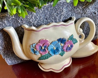 May include: Cream ceramic teapot-shaped planter with a scalloped rim and handle. It features blue and purple pansies and green leaves. The planter is set against a grey, textured background with green foliage.