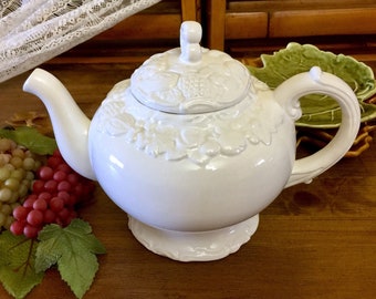 May include: White ceramic teapot with a grapevine design. The teapot has a lid and a handle. The teapot is sitting on a wooden surface.