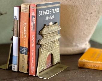 May include: A pair of gold-tone bookends shaped like a traditional Asian gate. The bookends are holding up three books, including two copies of Shakespeare plays, "Hamlet" and "Macbeth".
