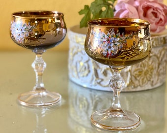 May include: Two gold-rimmed glass goblets with floral designs painted on the side. The goblets are on a white surface with a gold-trimmed box in the background.