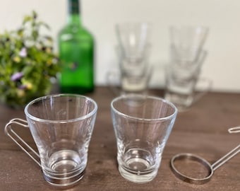 Op de afbeelding: Zes heldere glazen bekers met een metalen handvat aan twee van de bekers. De bekers staan op een houten oppervlak.