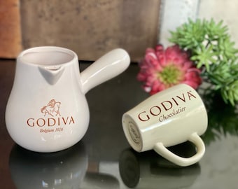 Könnte beinhalten: Ein weißer Keramik-Godiva-Heißgetränkekännchen mit Henkel und eine weiße Keramik-Godiva-Tasse mit dem Text "Godiva Chocolatier".