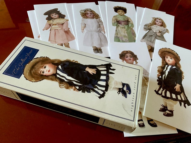 Puede incluir: Un conjunto de ocho tarjetas de mu&ntilde;ecas de estilo vintage que presentan diferentes mu&ntilde;ecas con diversos atuendos. Las tarjetas son blancas con un borde negro y presentan el texto "eighteen card set". Las tarjetas est&aacute;n dispuestas en forma de abanico sobre una superficie roja. Un libro titulado "The Golden Years" se encuentra en primer plano.
