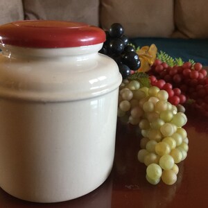 May include: A white ceramic jar with a red lid. The jar is sitting on a brown surface with a bunch of grapes in the background.