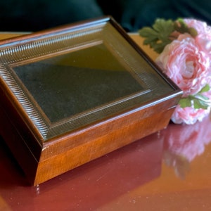May include: A wooden box with a glass top and a decorative border. The box is closed and sitting on a brown surface. A bouquet of pink flowers is next to the box.