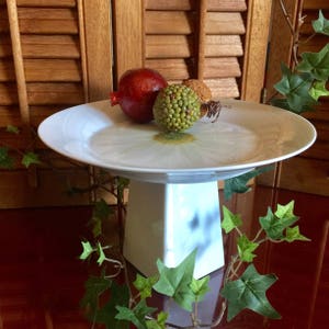 May include: White ceramic cake stand with a white daisy design. The stand has a round plate and a square base. There are three decorative fruit on the plate, one red and two green.