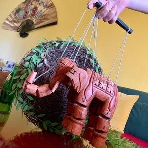 May include: A wooden elephant puppet with intricate carvings and a string control. The puppet is a warm brown color and is suspended in front of a decorative wreath. The background includes a decorative fan and a yellow wall.