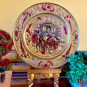 K&ouml;nnte beinhalten: Eine goldfarbene Metallwandplatte mit einem Reliefdesign von Personen in roten und goldenen Kleidern. Die Platte hat einen roten und goldenen Rand mit einem floralen Design. Die Platte steht auf einem Holzst&auml;nder mit drei Beinen.