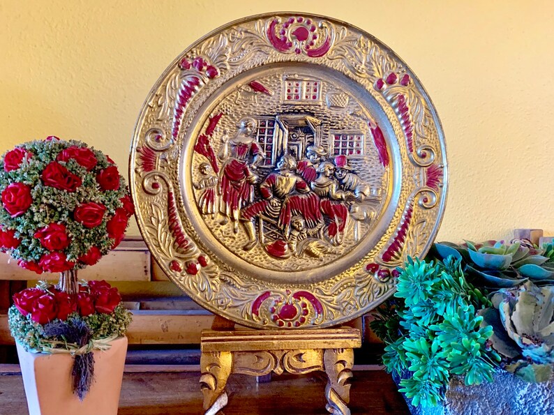 K&ouml;nnte beinhalten: Eine goldfarbene Metallwandplatte mit einem Reliefdesign von Personen in roten und goldenen Kleidern. Die Platte hat einen roten und goldenen Rand mit einem floralen Design. Die Platte steht auf einem Holzst&auml;nder mit drei Beinen.