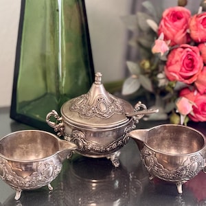 K&ouml;nnte beinhalten: Ein silbernes Zucker- und Sahne-Set mit verzierten Designs. Die Zuckerdose hat einen Deckel und einen kleinen L&ouml;ffel. Die Kanne hat einen Griff und einen Ausguss. Das Set befindet sich auf einer dunklen Oberfl&auml;che.