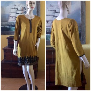 May include: A mustard yellow tunic dress with black embroidery around the neckline and hem. The dress has three-quarter sleeves and a relaxed fit.