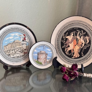 May include: Three decorative plates with Greek-themed designs. The largest plate features a black border and a central image of figures. The other plates depict Greek architecture and figures in a circular design. The plates are displayed on black stands.
